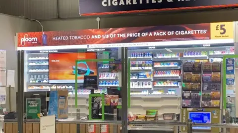 Kios tembakau di toko Morrisons yang menunjukkan iklan untuk dua merek tembakau yang dipanaskan, piring dan IQOS, di belakang layar Perspex. Slogan Ploom membaca "Tembakau yang dipanaskan dan 1/3 harga sebungkus rokok"