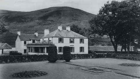 Tweedsmuir Community Company An old black and white picture of the Crook Inn in Tweedsmuir - a white building in among trees with hills in the background