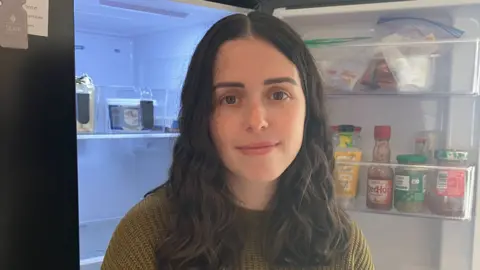 A lady with shoulder length brown hair smiles as she wears a green jumper. She is standing in front of an open fridge.
