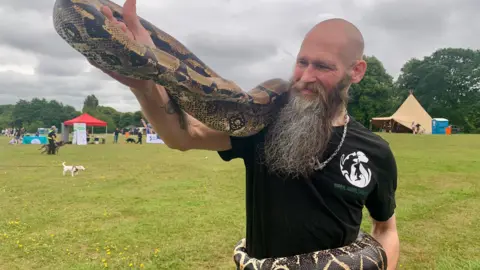 Dave Copson-Moore of the Wirral Animal Samaritans with a shaved head and long brown and grey beard and black t-shirt standing in a park holding a boa constrictor snake which is wrapped around his waist and arm.