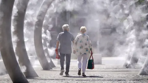 A man and a woman - both with silver hair - are walking inside a row of ring metal structures that are spraying mist on a very sunny day. A man is wearing a checkered blue shirt with short sleeves, jeans and trainers and a woman in white trousers and a loose-fitting shirt.   