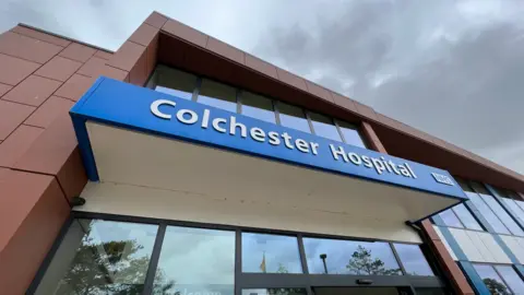 A red block two-storey building with glass facade, made up of windows with black frames. There is a portico above the entrance with the words Colchester Hospital in white letters on a blue background. Trees are reflected in the windows and the sky has dark clouds.