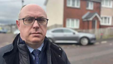 A man with glasses standing outside across the street from a terraced house. The house and car outside it are out of focus in the background. The man is wearing a grey coat, navy top, blue shirt and navy tie.