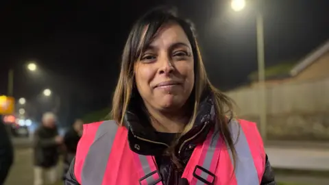 Bea Lane has long brown hair and is wearing a pink high-vis jacket. She is standing on a pavement with street lights in the background.