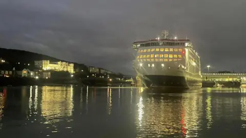 The Manxman, which is a large red, white, and black ferry, sitting in low water in Douglas Harbour at night. The lights from the ship, the harbourside buildings are reflecting on the water around it.