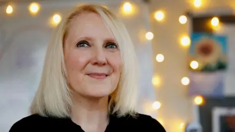 Tressa Burke, who has shoulder length blonde hair, looks off into the distance. There are lights and a picture of a sunflower on the wall behind her.
