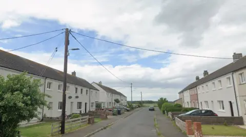 A street with houses on either side. There is a green field at the end of the road. 