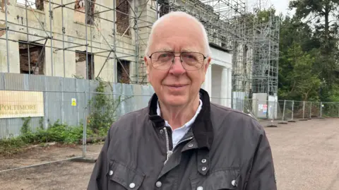 A grey haired man , wearing glasses and a dark jacket, stood outside the remains of a mansion house, surrounded by scaffolding.