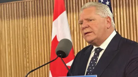 Doug Ford speaks into a mic, wearing a black suit with a blue tie.