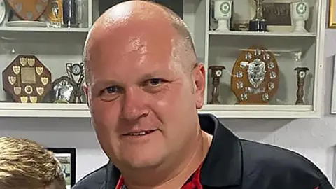 Head and shoulders shot of a bald man wearing a dark polio shirt in front of a trophy cabinet wall.