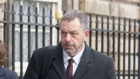 BBC Tony Chambers, who has short grey hair and a grey beard, walks towards Liverpool Town Hall wearing a dark grey suit and sets his face in a serious expression