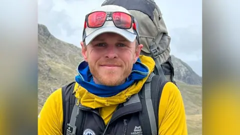 Alex Jones A head-and-shoulder image of Alex Jones on a mountain. He has a trimmed ginger beard and blue eyes. He is wearing a blue and yellow outdoor jacket, a grey baseball cap with red-tinted sunglasses perched on top and a large rucksack. Behind him are the tops of craggy hills.