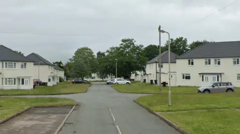 A Google Streetview image of a residential street. A number of houses, all white with grey roofs, are on each side of the road and cars are parked at various points.