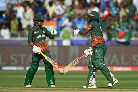 Jewel Samad/AFP Two cricketers dressed in green prepare to fist bump while holding their cricket bats. There is a crowd of spectators behind them.