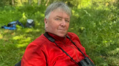 Aidan Crean wears a red fleece and has a pair of binoculars round his neck. He has grey hair and a neutral expression. 