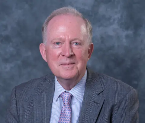 David Chambers David Chambers wearing a grey suit with a pink and purple tie, looking at the camera against a grey backdrop.