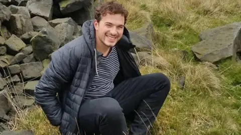 Family handout Jack Ritchie, a man in his 20s with dark hair and wearing black jeans, a striped T-shirt and a black jacket, sitting down in a field next to a stone wall.