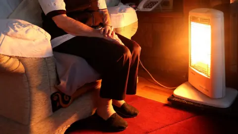 An elderly person wearing a white top under dark clothing sits next to a glowing bar fire on a red carpet with their arms folded on their lap.