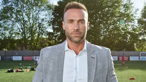 Best id wearing a grey suit and white shirt and standing on a football field in the summer sun. He has brown spiky hair and a close-cropped beard.
