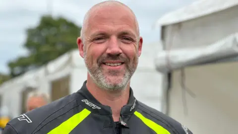 John Eastham has short grey hair and a beard, he wears a black jacket with a fluorescent yellow stripe. 