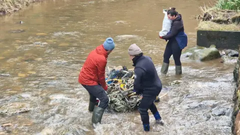 This picture shows volunteers from British Divers Marine Life Rescue clearing plastic waste from Boggle Hole, near Robin Hood's Bay. Two volunteers, stand ankle-deep in the water dragging a large bundle of litter. A third volunteer stands behind them, holding a bag of waste.