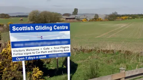 A rural road runs alongside open fields under a clear blue sky, with a sign for the Scottish Gliding Centre visible near a wooden fence.