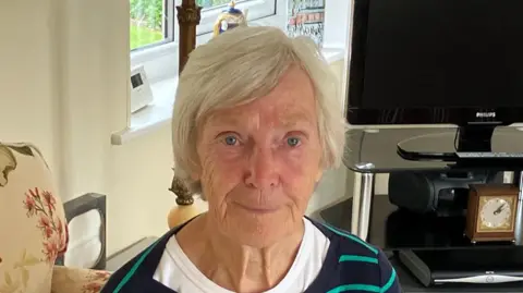 Veronica Bird. She has short blonde hair and sits in a living room. She is wearing a white T shirt and stripey jumper.