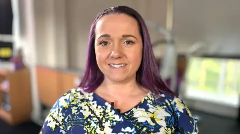 Martin Giles/BBC Angel Clark is wearing a blue dress with white and yellow flowers and black stems. She has purple hair and is smiling at the camera. 
