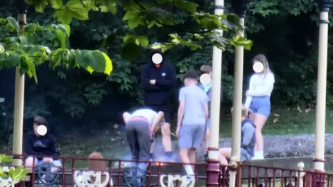 A group of six boys and one girl with blurred faces stand along a fire near a park bandstand. Smoke is rising from the small fire. 