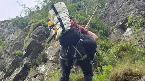 Patterdale Mountain Rescue A mountain rescue volunteer using ropes to rescue a dog at Buck Crag, Hartsop. The volunteer is pictured from behind, climbing back up the rock face. They are carrying a black staffy-type dog under their arm and are carrying a large rucksack on their back.