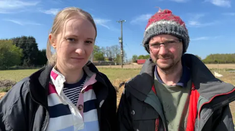 A woman and a man stand in front of a green field under a blue sky. The woman has blond hair tied back and is wearing a rugby shirt with blue, white and pink stripes and a black jacket. The man is wearing a grey and red bobble hat, glasses, a grey coat and a green jumper.