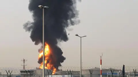 Stringer/EPA/Shutterstock Smoke rises from an area near Dubai International Airport in Dubai, United Arab Emirates amid the US-Israel conflict with Iran. Spiralled barbed wire on Y-shaped props surrounds the perimeter with concrete buildings beyond. In the foreground is a red and white striped pole. To the left is a smokey ball of fire with a large plume of black smoke rising from it.