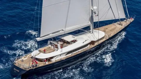 PERINI NAVI PRESS OFFICE The Bayesian yacht with the lower parts of its white sails visible above the whole of its brown deck. The cabins have white roofs and the hull of the boat is black. About half a dozen people in red tops are visible on deck.