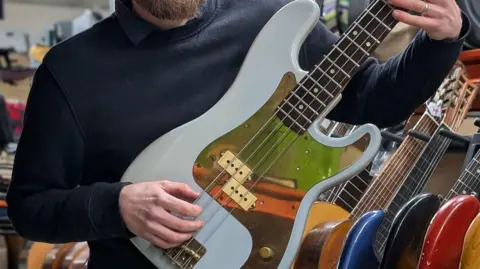 PA Media A man wearing a navy blue jumper holding a bass guitar. The guitar is white with a gold inlay.
