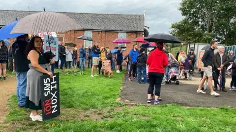 Dozens of residents stand in the rain at a protest. Some are holding placards and signs opposing the plans for the clinical waste incinerator. Other residents are holding umbrellas.