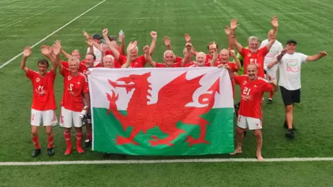BBC A football team featuring players all over the age of 70. They are holding a Welsh flag with their arms in the air celebrating victory. They are stood on a football pitch with the lines visible. Some have football bots on whereas others have socks or are barefoot.