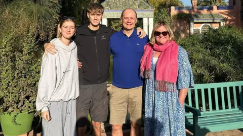 Family Four people stand in a line on a sunny day, their arms around each other. A young woman on the left is wearing a light grey hoodie and darker grey tracksuit trousers. Next to her is the young man from the above picture, wearing a black hoodie and grey shorts. On his left is a middle-aged man wearing a blue polo shirt and light brown shorts, and a middle-aged woman wearing sunglasses, a blue and white dress and a pink pashmena.