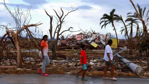 Reuters A young woman, boy and an adult walk past widespread debris and damaged trees. A mound of collapsed housing is in the background.