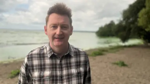 BBC Neil Reid, who has spiky brown hair and a checked shirt stands in front of a green lough