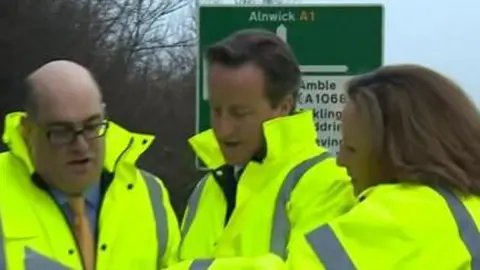 David Cameron, the then MP for Berwick, Anne-Marie Trevelyan and another politician all wearing hi-vis jackets stand in front of a sign pointing to Alnwick 