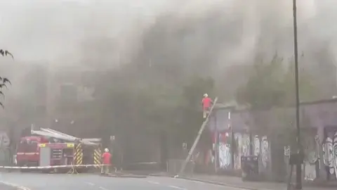 Fire fighters tackle a blaze amid thick black smoke at a derelict building in Manchester city centre with one firefighter on top of a ladder.