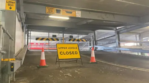 LDRS A upper level closed sign sits in the middle of a ramp leading up to further floors in a car park. Bollards are there too.