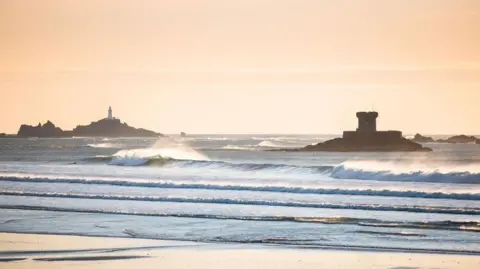 Andy Le Gresley Photography A coastal scene at sunset or sunrise with soft golden light. In the foreground, there is a sandy beach with shallow water and gentle waves rolling in. Further out, you can see the sea with multiple lines of breaking waves.
In the mid-ground, there is a stone tower on a rocky outcrop, likely a historic coastal fortification or defensive structure