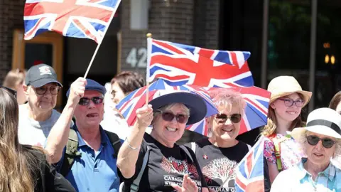 AFP/Getty Images Well wishers wave flags before the arrival of Britain's King Charles III and Queen Camilla at a Lansdowne Park community event in Ottawa, Canada, May 26, 2025.