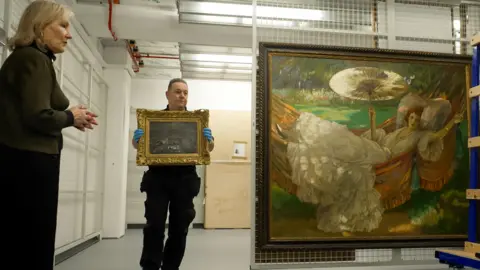 A man carrying a painting in a gold frame into a large room. He is wearing blue protective gloves. A woman is standing to the left of the picture, she is looking at a large painting of a woman lying in a hammock 