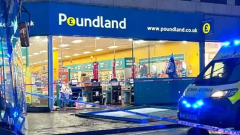 Tom Whiting The front of a Poundland store with a broken window lying in the street and an ambulance parked outside, with blue and white police tape around it