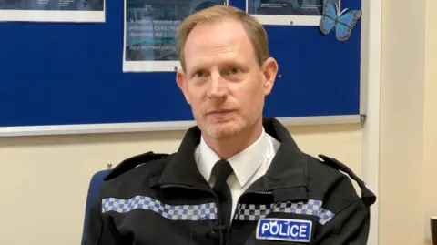 Chief Superintendent Simon Dodds has short light brown hair. He is wearing police uniforn and is sat at a desk.