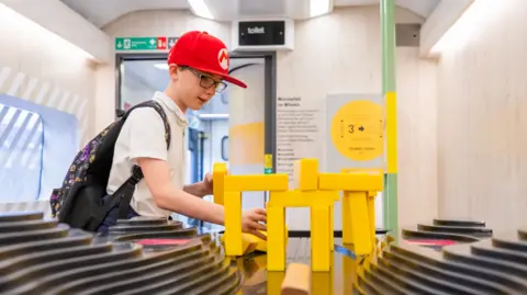 Jack Boskett/Railway 200 A boy of about 11, standing in a converted train carriage, wearing a white polo shirt and red baseball cap with a rucksack on is back and handling an exhibit of yellow bricks, which he is making into a bridge