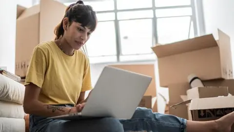 Getty Images Uma jovem olha para um laptop cercado por caixas de papelão