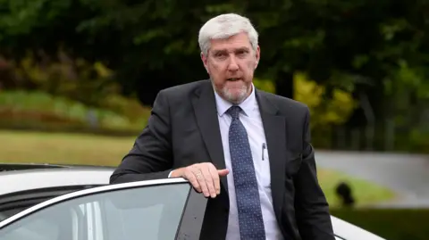 Mark Marlow/PA John O'Dowd gets out of a car. He has short grey hair and a grey/ginger beard and is wearing a dark suit and tie and a light blue shirt